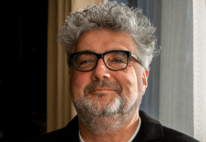 Smiling middle-aged man with gray hair and beard, wearing black-framed glasses, indoors near a window.