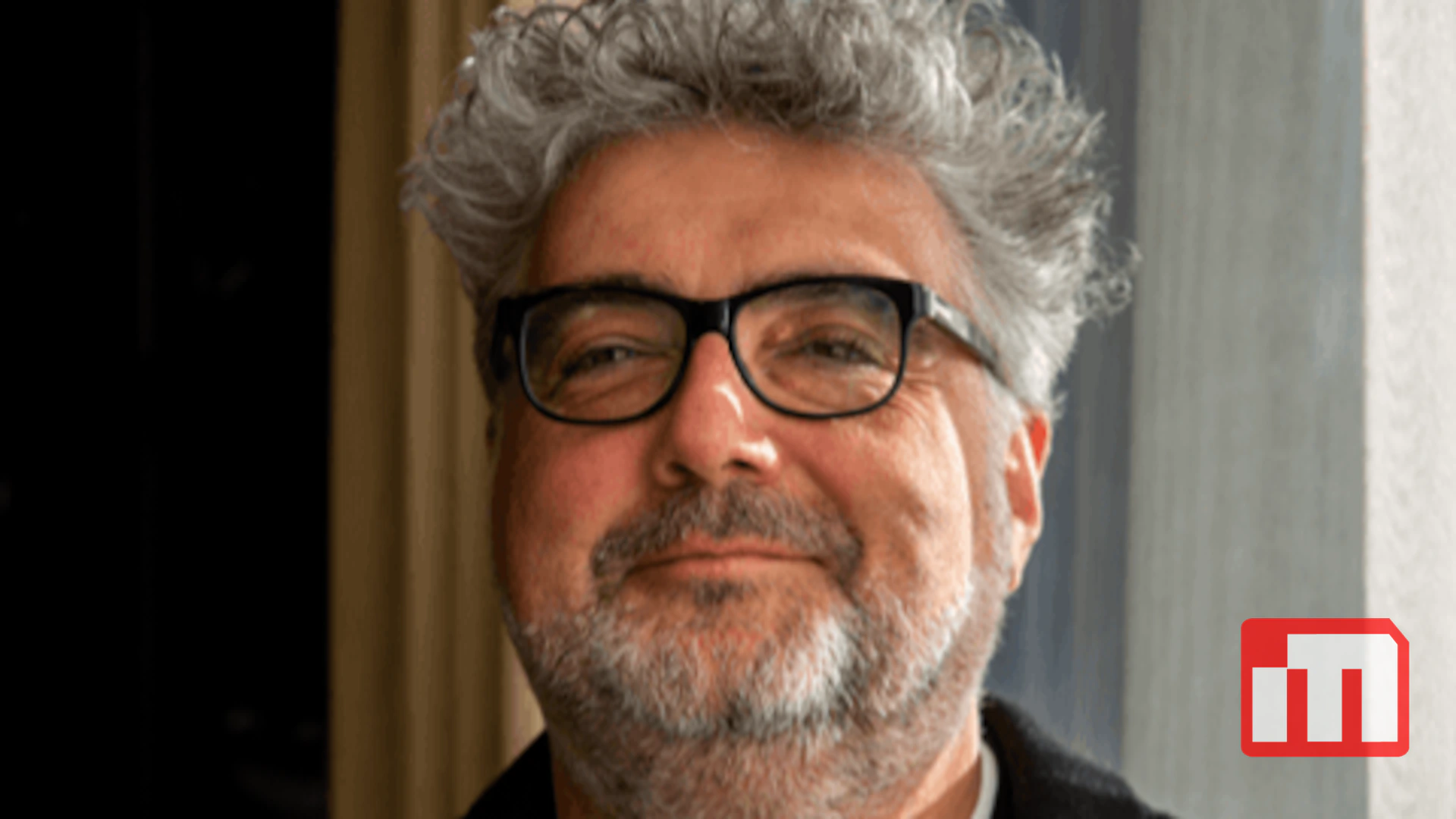 Smiling middle-aged man with gray hair and beard, wearing black-framed glasses, indoors near a window.