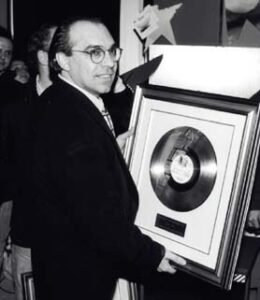 Man in a suit and glasses holding a framed platinum record award at an event.