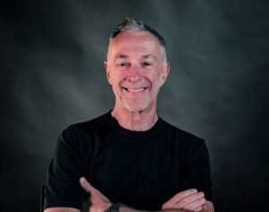 Smiling man with crossed arms wearing a black shirt against a dark studio backdrop.