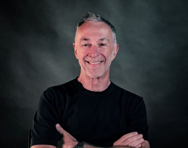 Smiling man with crossed arms wearing a black shirt against a dark studio backdrop.