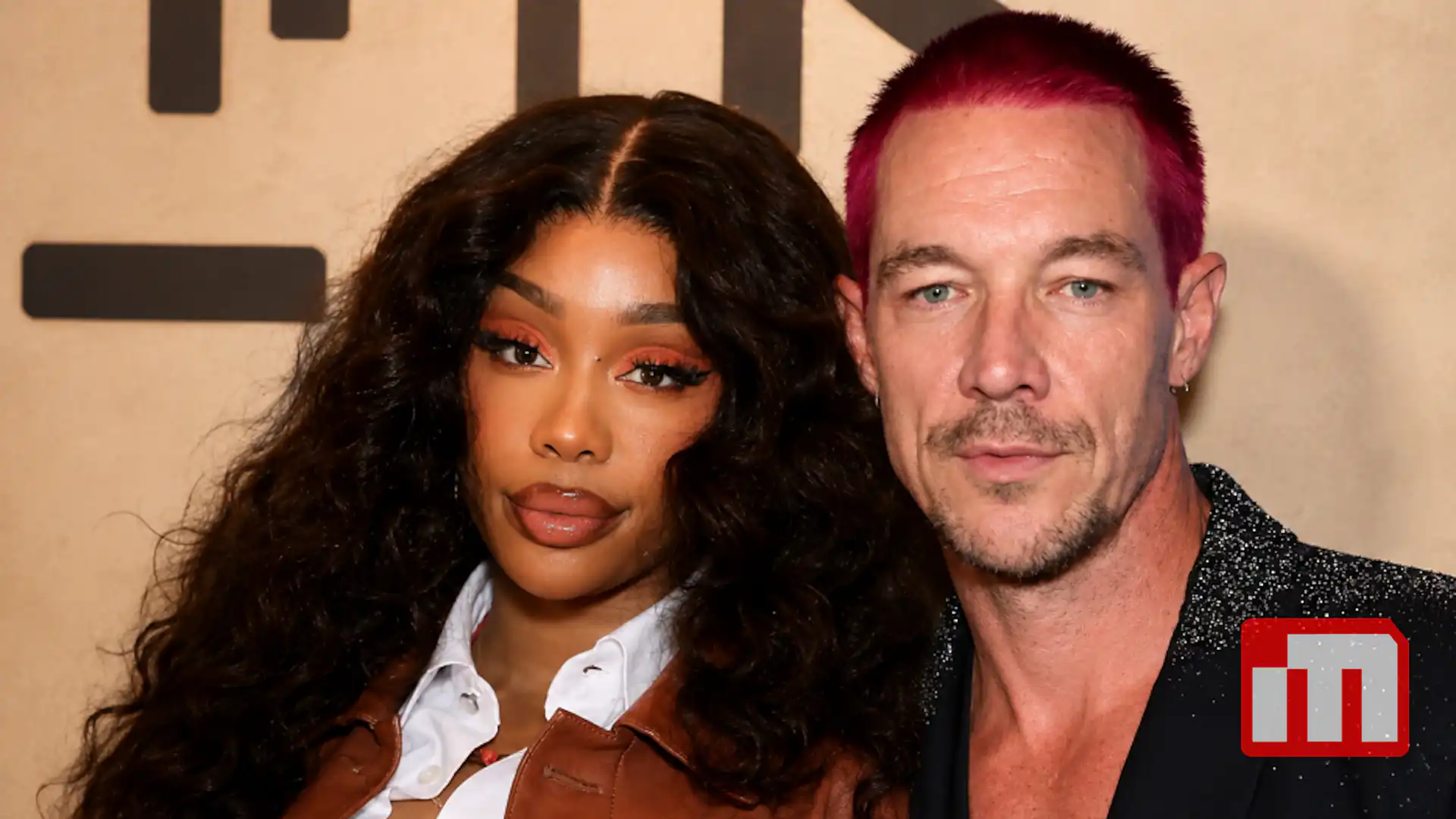 Woman with long dark curls wearing a white shirt and brown leather jacket standing next to a man with bright pink hair in a black glittery suit at a red-carpet event.