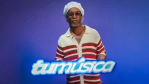 Man in a white headwrap and sunglasses stands against a blue backdrop, with a glowing 'tumusica' sign in front.