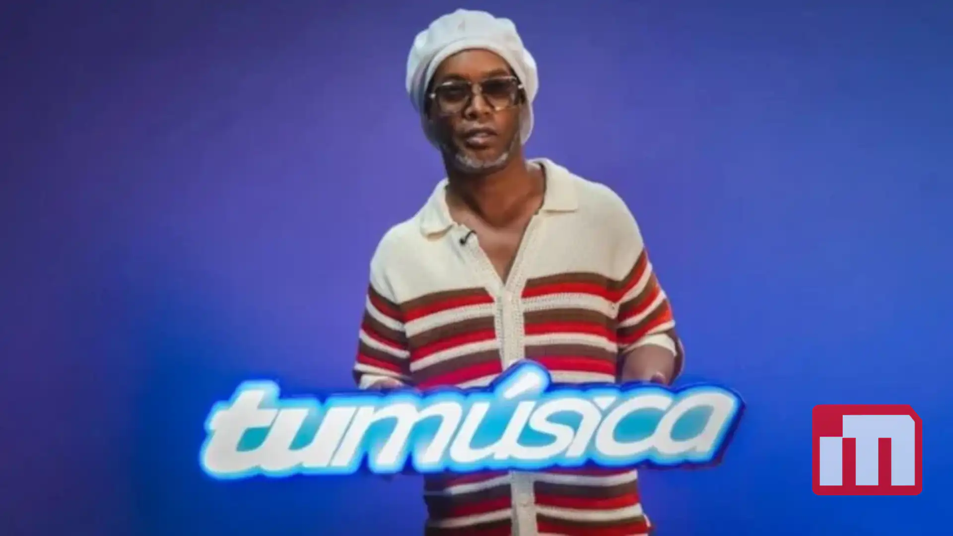 Man in a white headwrap and sunglasses stands against a blue backdrop, with a glowing 'tumusica' sign in front.