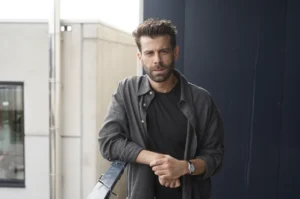 Bearded man in a black T-shirt and gray shirt stands on a rooftop balcony, leaning on a railing.