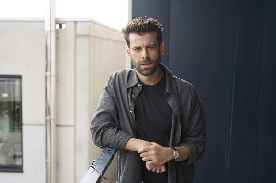 Bearded man in a black T-shirt and gray shirt stands on a rooftop balcony, leaning on a railing.